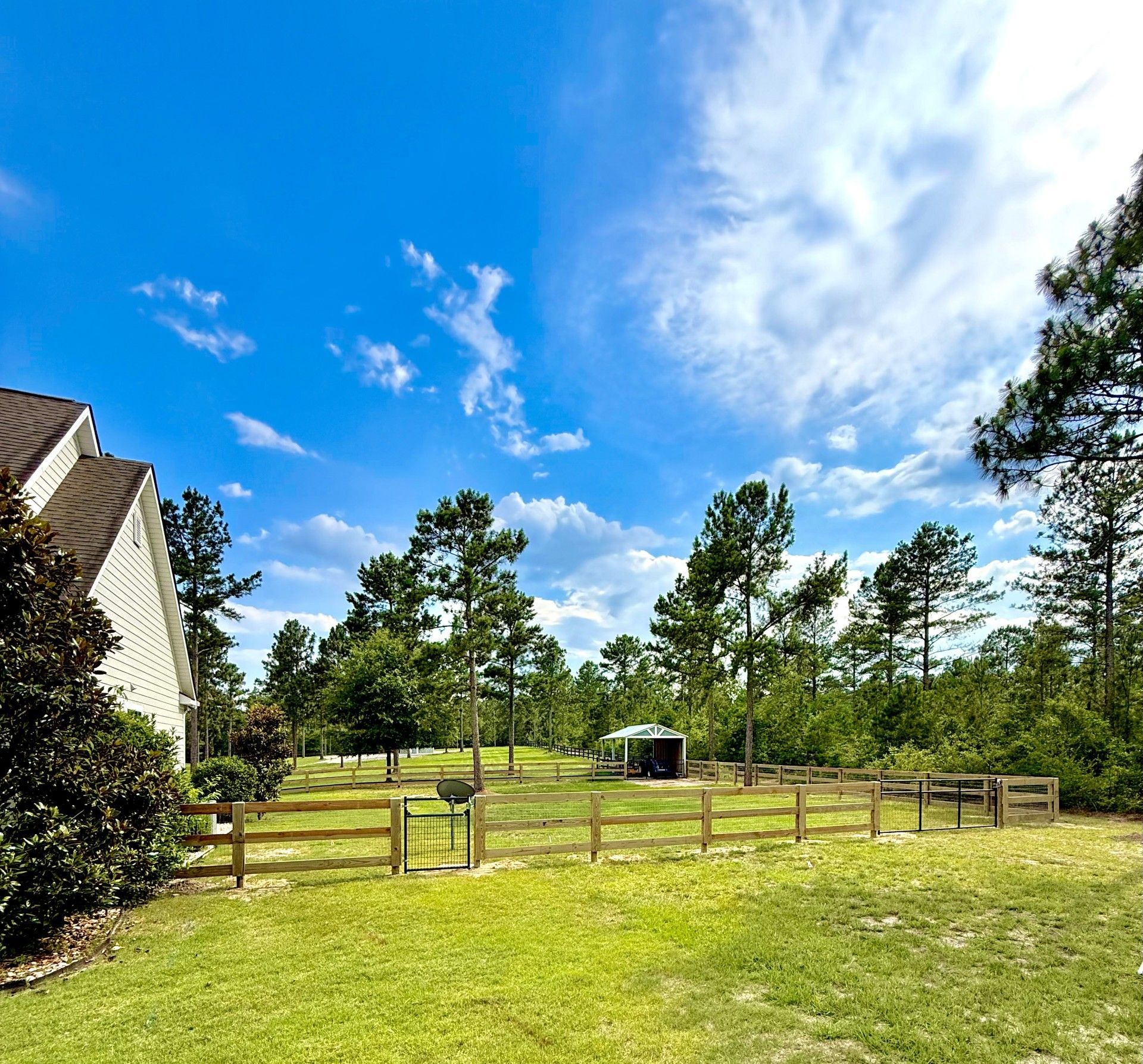Wooden fence beside a dirt path and grassy field with trees under a clear blue sky.