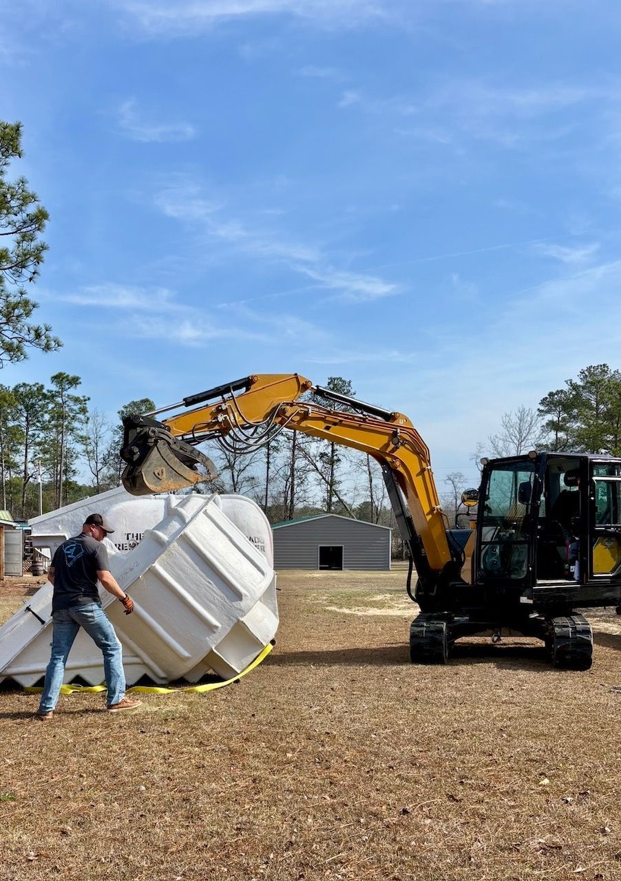 Man guiding a yellow excavator tipping a large white tank outdoors on wood chips under a blue sky