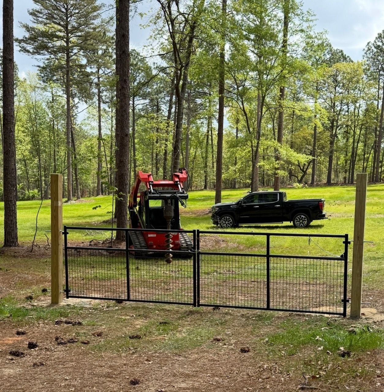Red skid steer and black pickup truck by a black metal gate in a wooded yard.