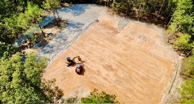 Aerial view of two people working on a large cleared dirt lot surrounded by trees and water.