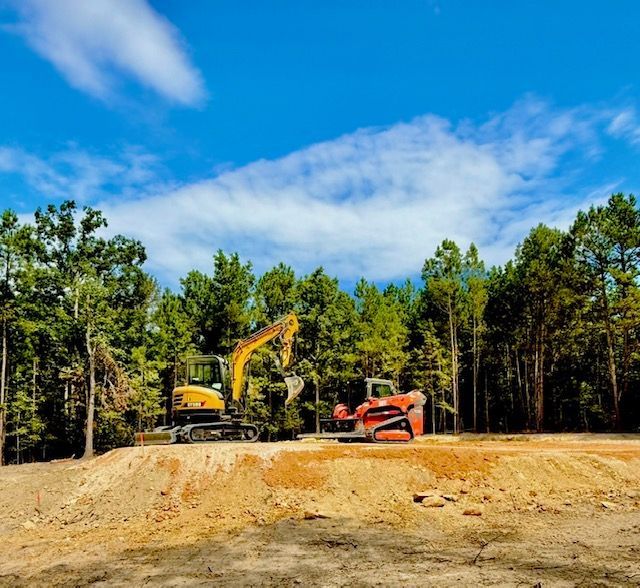Yellow excavator and orange bulldozer on a dirt site under a blue sky, surrounded by trees