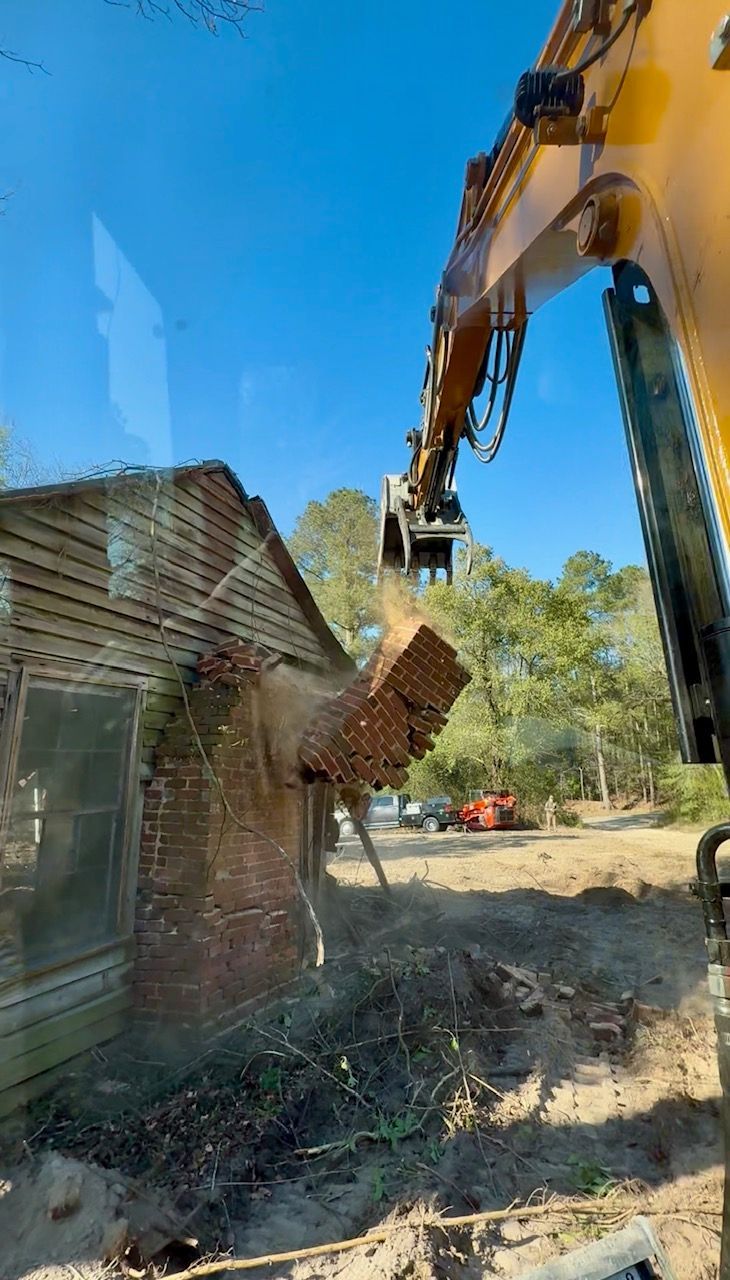 Excavator demolishing a brick wall beside a house on a sunny day