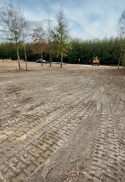 Tire-tracked dirt lot beside trees, with a small construction vehicle in the distance.
