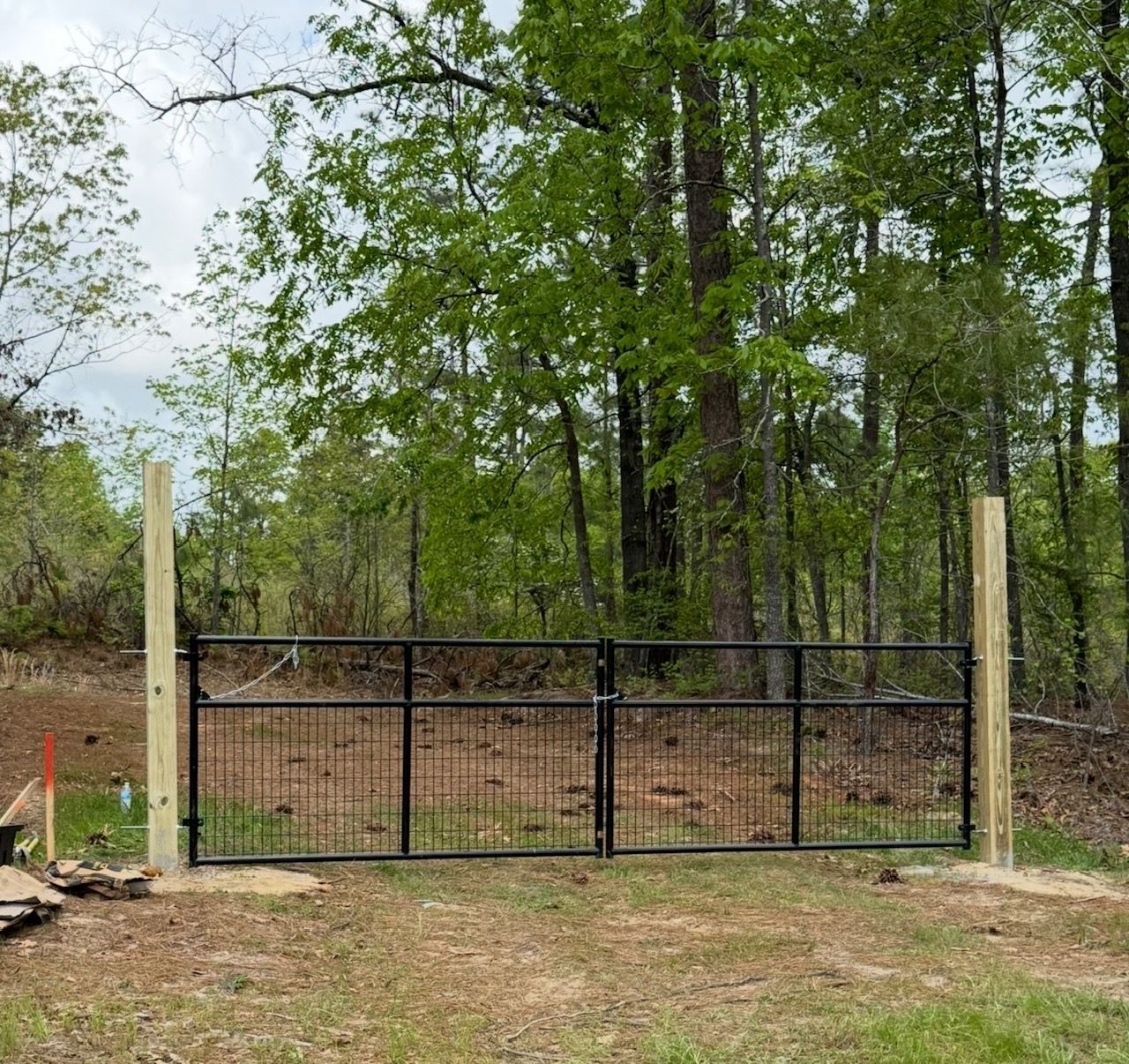Black metal driveway gate between tall wooden posts in a wooded yard