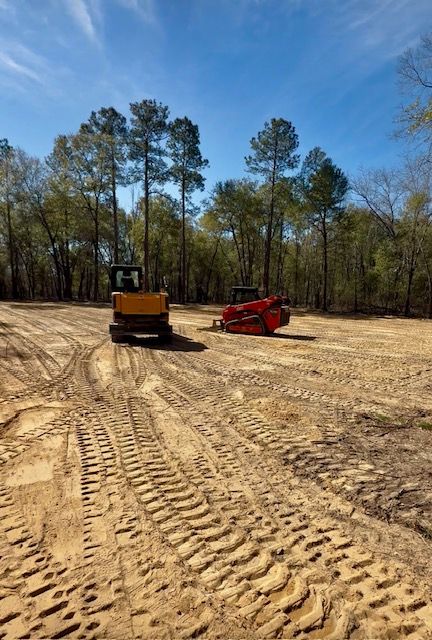 Bulldozer and red compact loader on a dirt clearing with tire tracks and trees under a blue sky