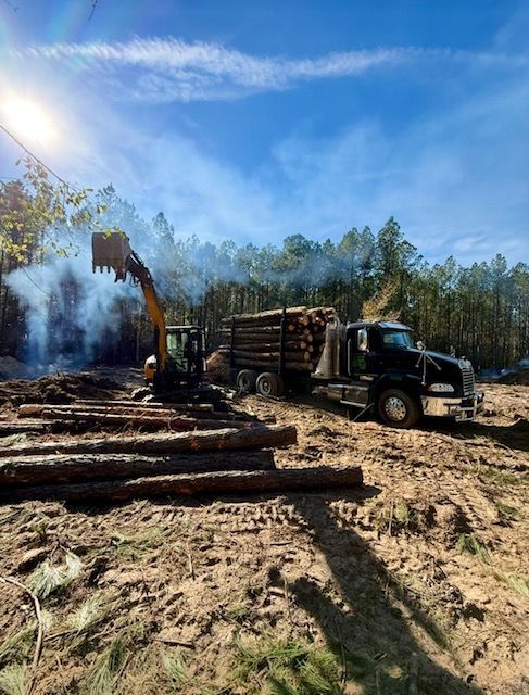 Logging truck loaded with cut timber beside a skidder in a smoky forest clearing under blue sky