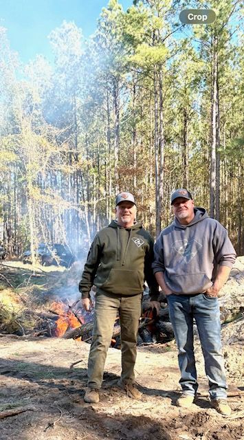 Two people standing by a smoky forest fire, with tall trees and flames in the background.