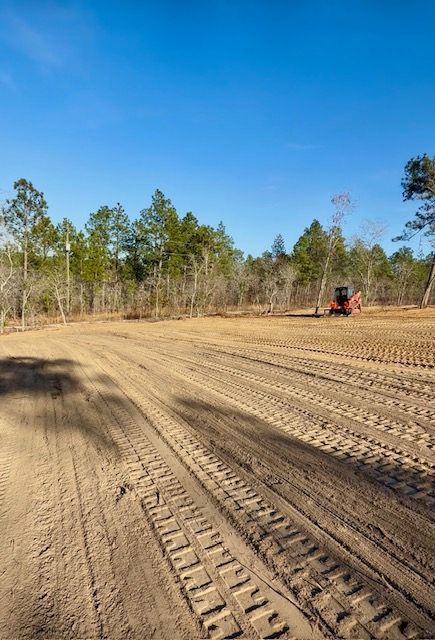 Wide sandy dirt lot with tire tracks, trees along the edge, and a small red tractor in the distance.