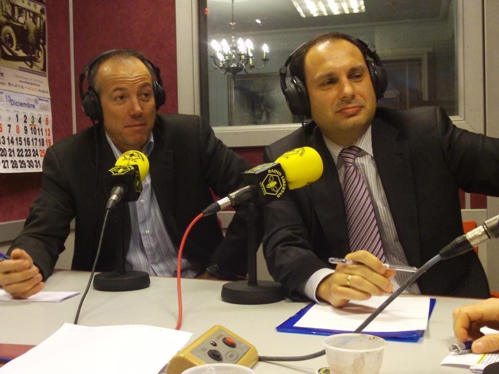 Dos hombres de traje que usan auriculares en un estudio de radio y hablan a través de micrófonos.