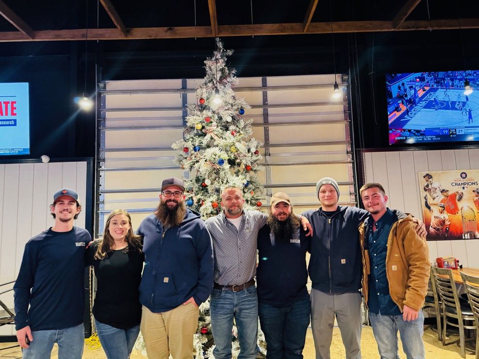Group posing indoors in front of a decorated Christmas tree, with TVs and restaurant seating nearby. Group posing indoors in front of a decorated Christmas tree, with TVs and restaurant seating nearby.