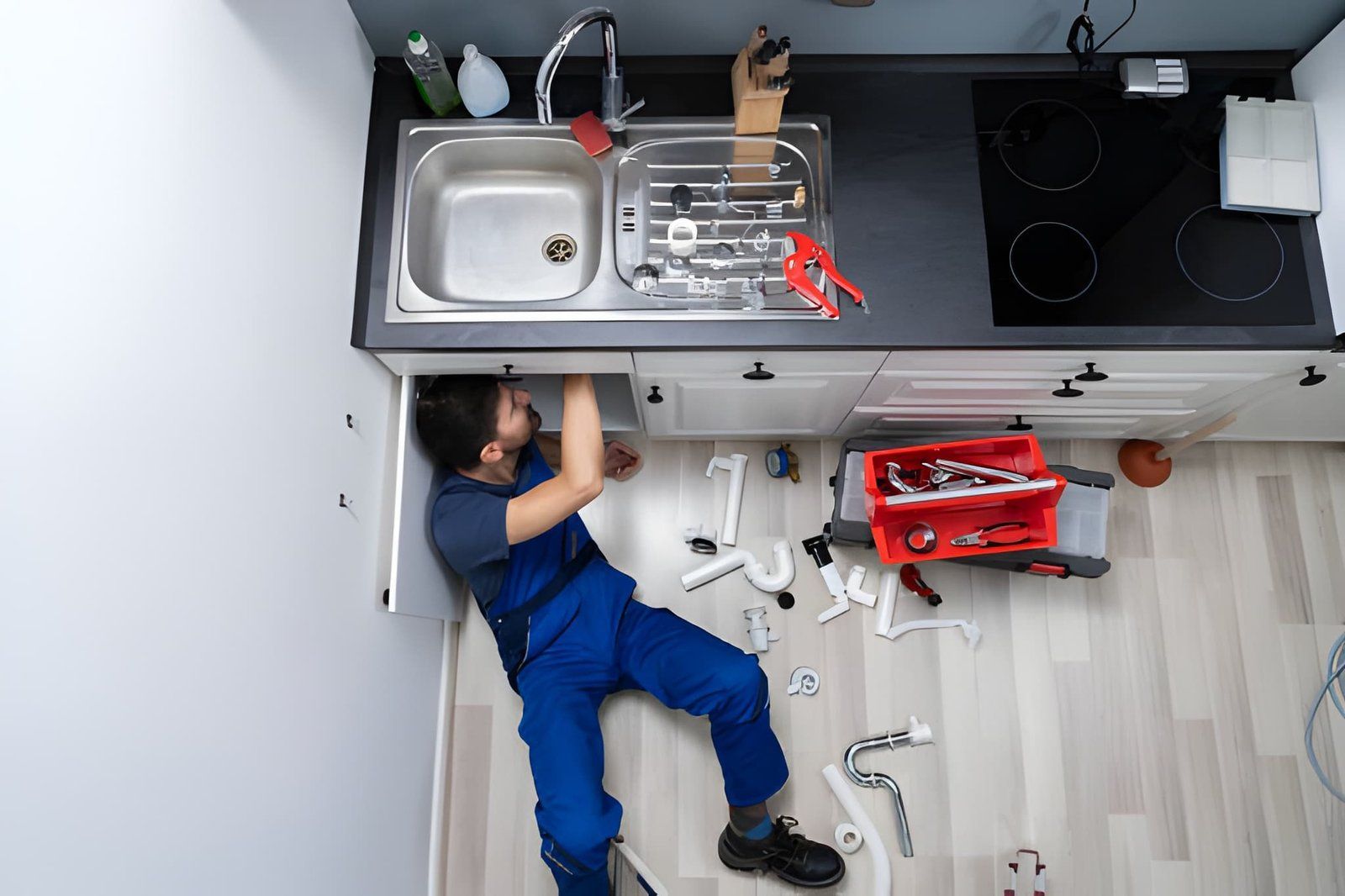Technician in blue uniform lies on floor under kitchen counter, working near sink and tools.
