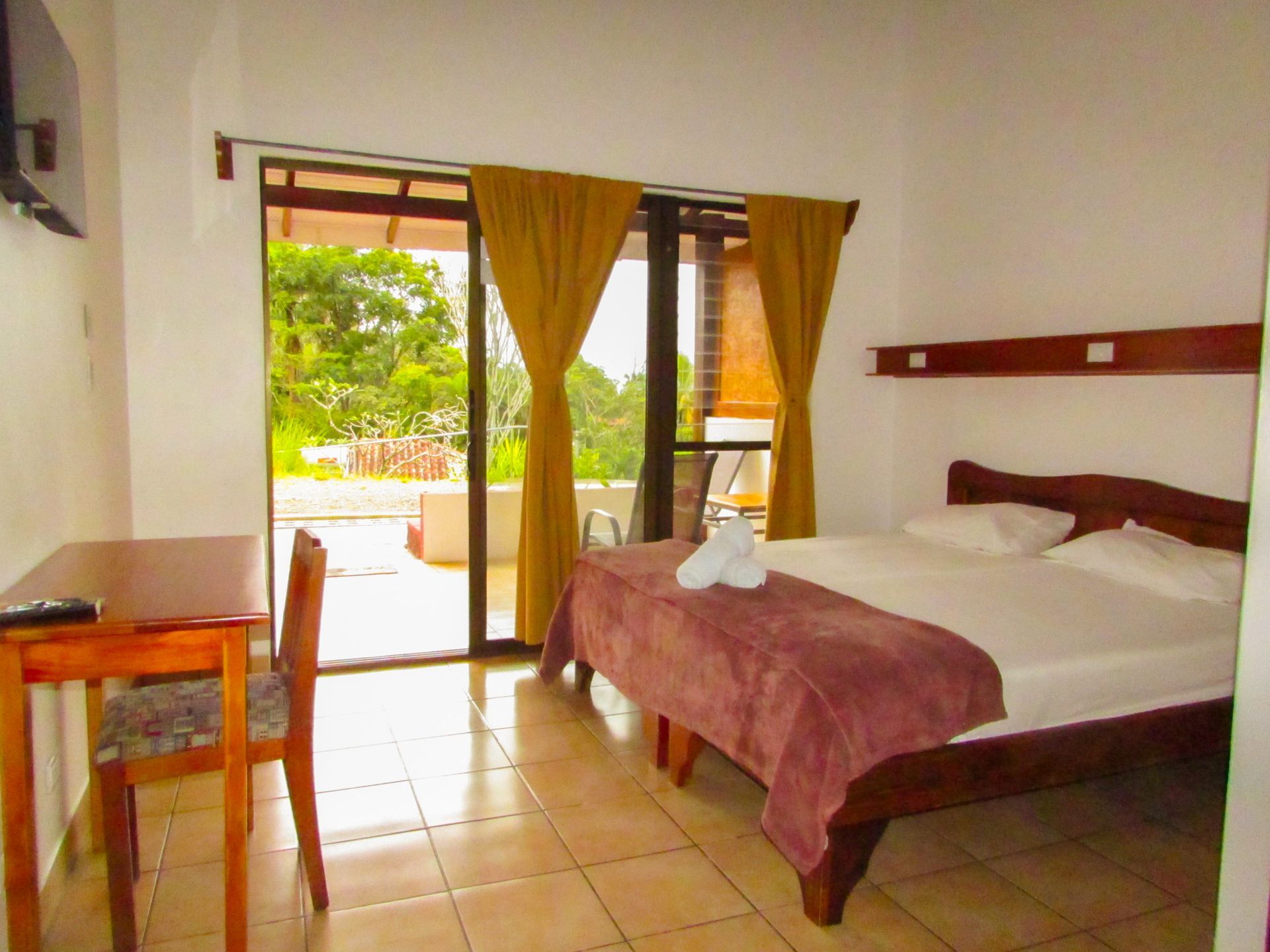 Habitación con cama doble, mesa de madera y puertas correderas que dan a un balcón con vegetación.