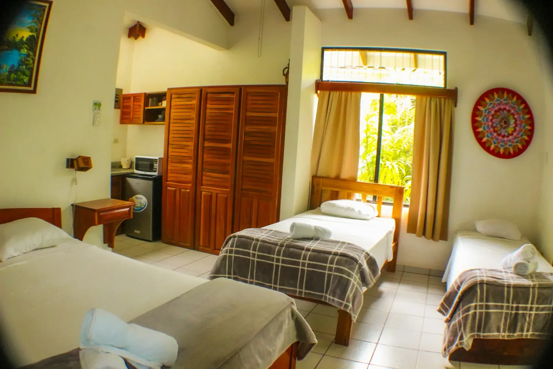 Habitación de hotel sencilla con tres camas, armario de madera y luz natural que entra por la ventana.