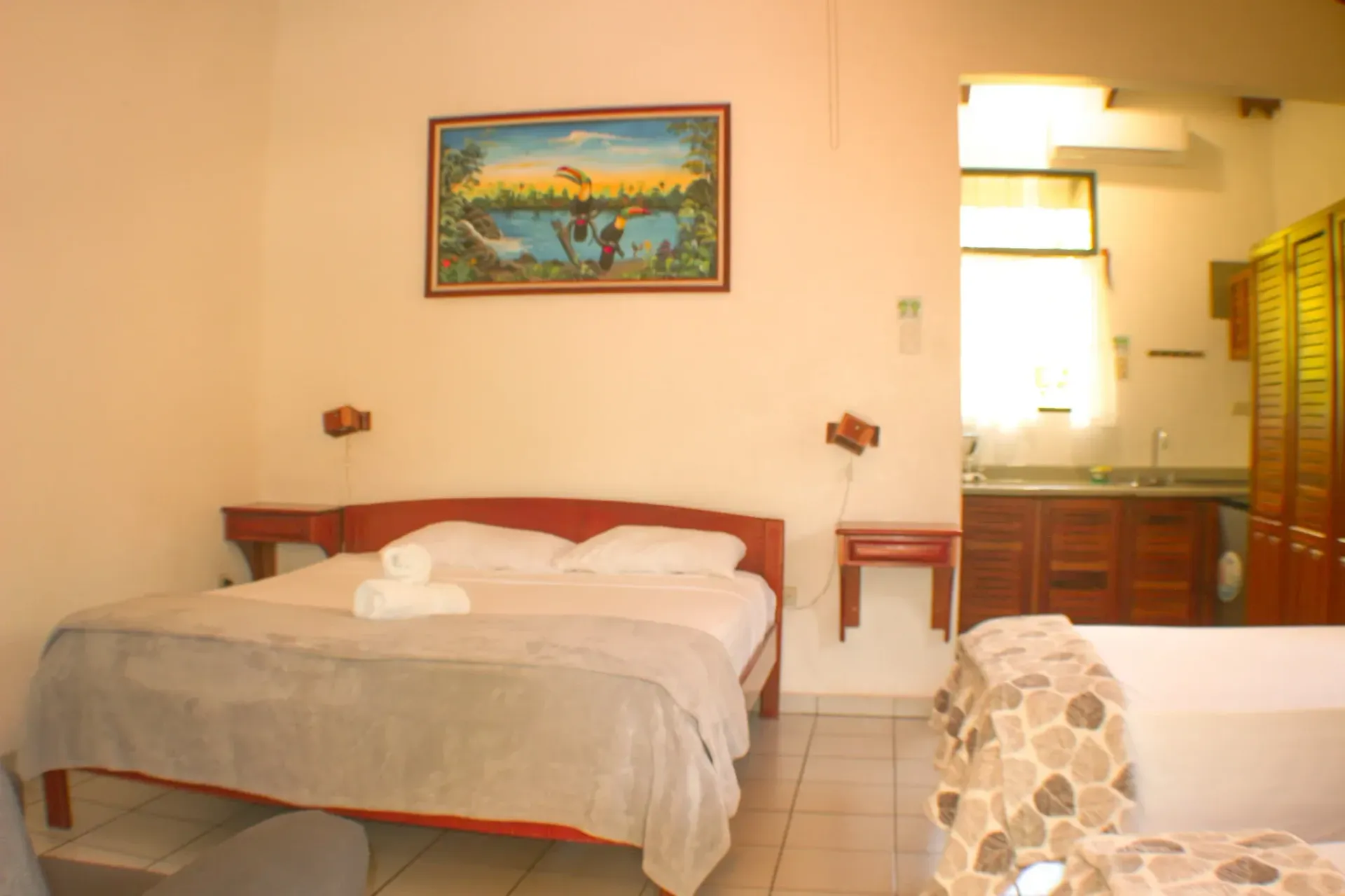 Habitación de hotel pequeña con cama, cuadros en la pared, mesitas de noche y vistas al baño y a la zona de cocina.