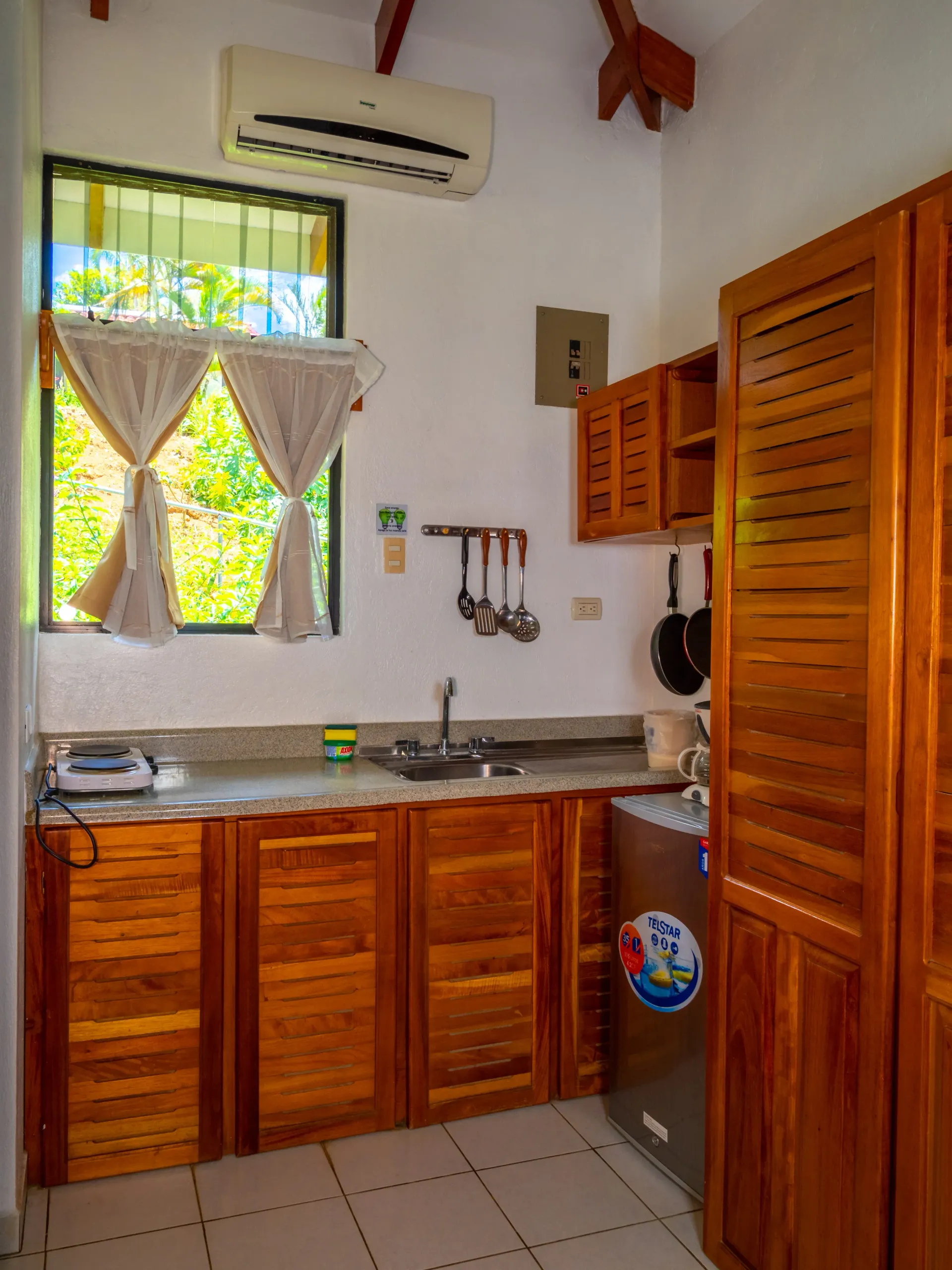 Cocina pequeña con armarios de madera, fregadero de acero inoxidable, suelo de baldosas, ventana y aire acondicionado mural.
