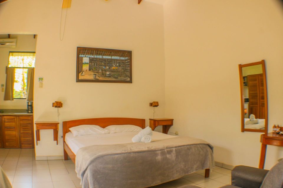 Habitación de hotel sencilla con cama, mesita, espejo y paredes de color beige cálido.