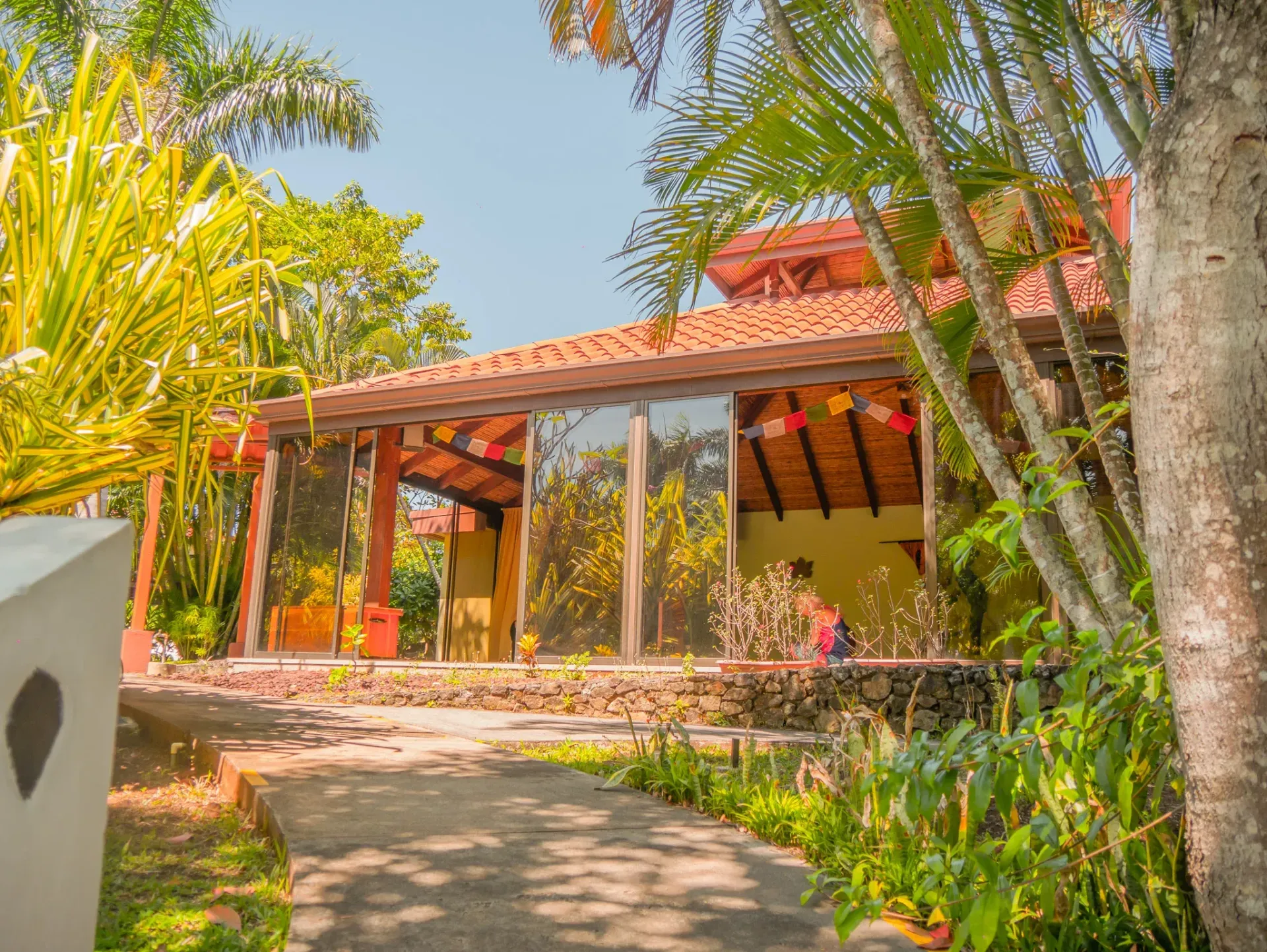 Sunlit tropical house with red tile roof, palm trees, and a garden path leading to the porch