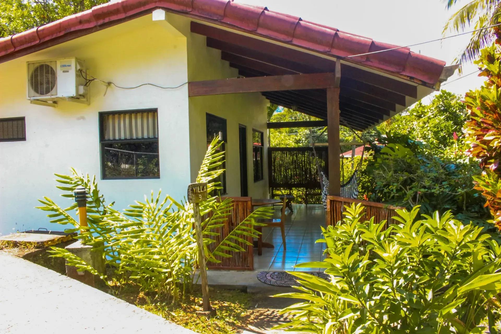 Small white house with a red roof, surrounded by lush green plants and a shaded porch.