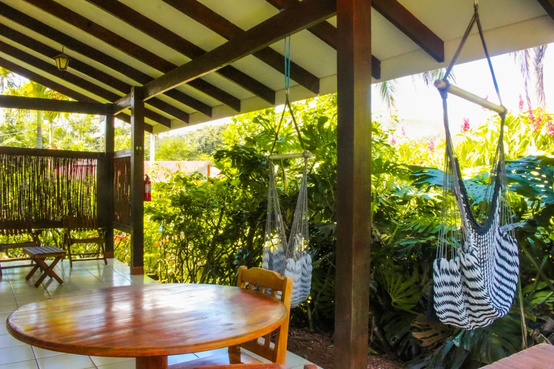 Patio cubierto con mesa de madera, silla colgante a rayas y vistas a un exuberante jardín verde.