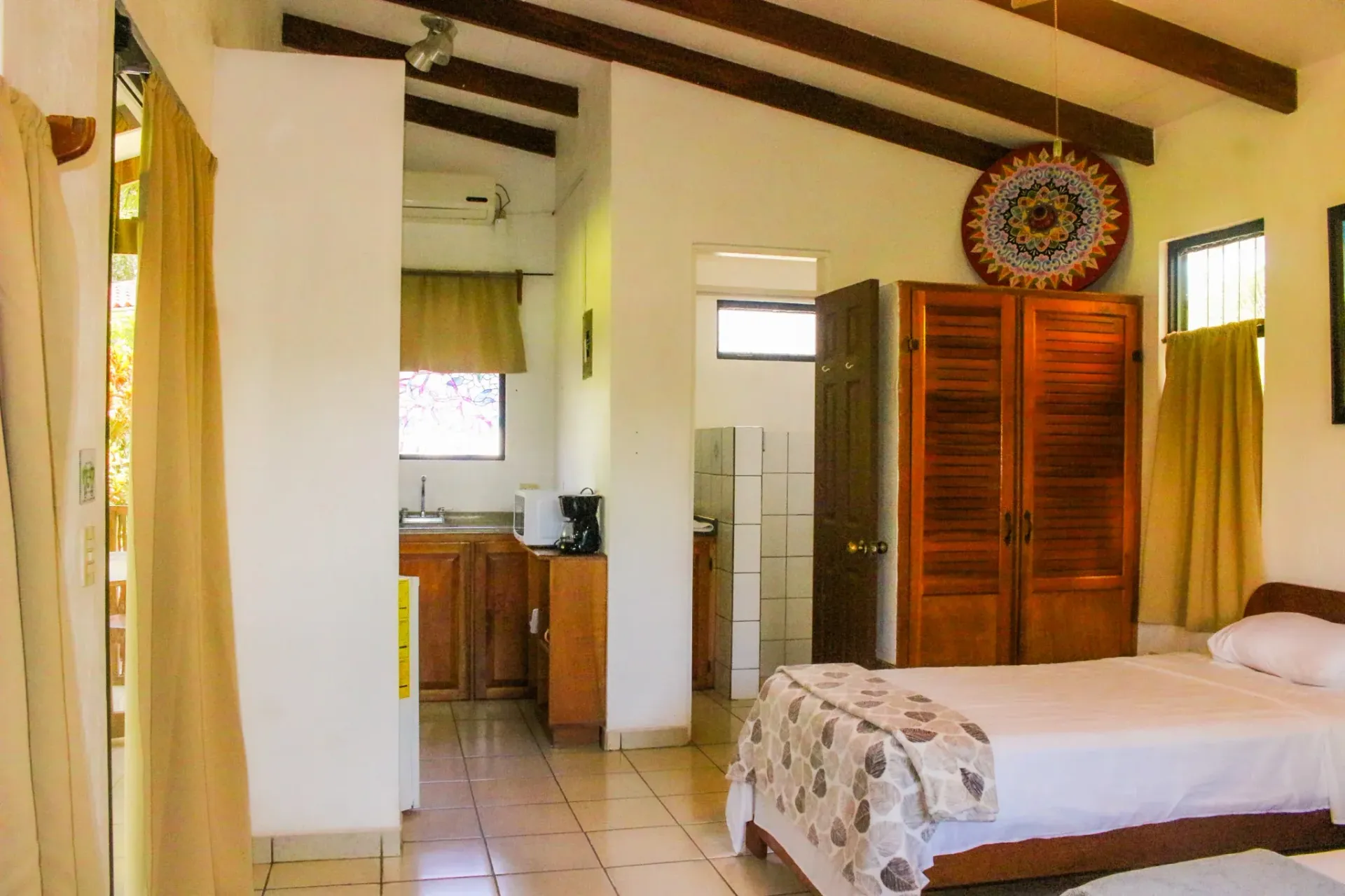 Habitación luminosa con cama, armario de madera, suelo de baldosas y una pequeña zona de cocina en la parte trasera.