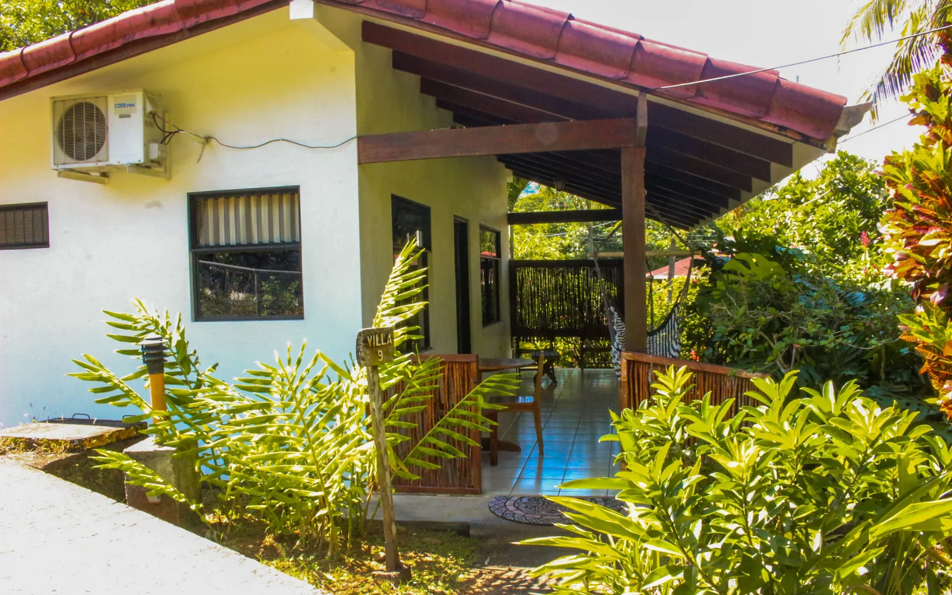 Pequeña casa blanca con techo rojo, porche y exuberantes plantas tropicales en primer plano.
