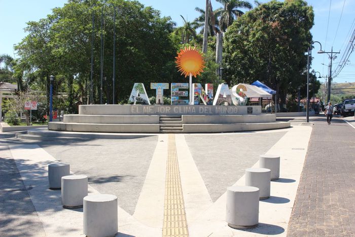 Letrero de avenida con el logotipo de un sol en una plaza, flanqueado por árboles y bolardos de hormigón