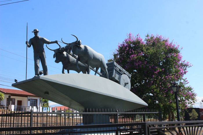 Estatua de un vaquero y un toro sobre una plataforma inclinada, con un árbol en flor y un cielo azul detrás.