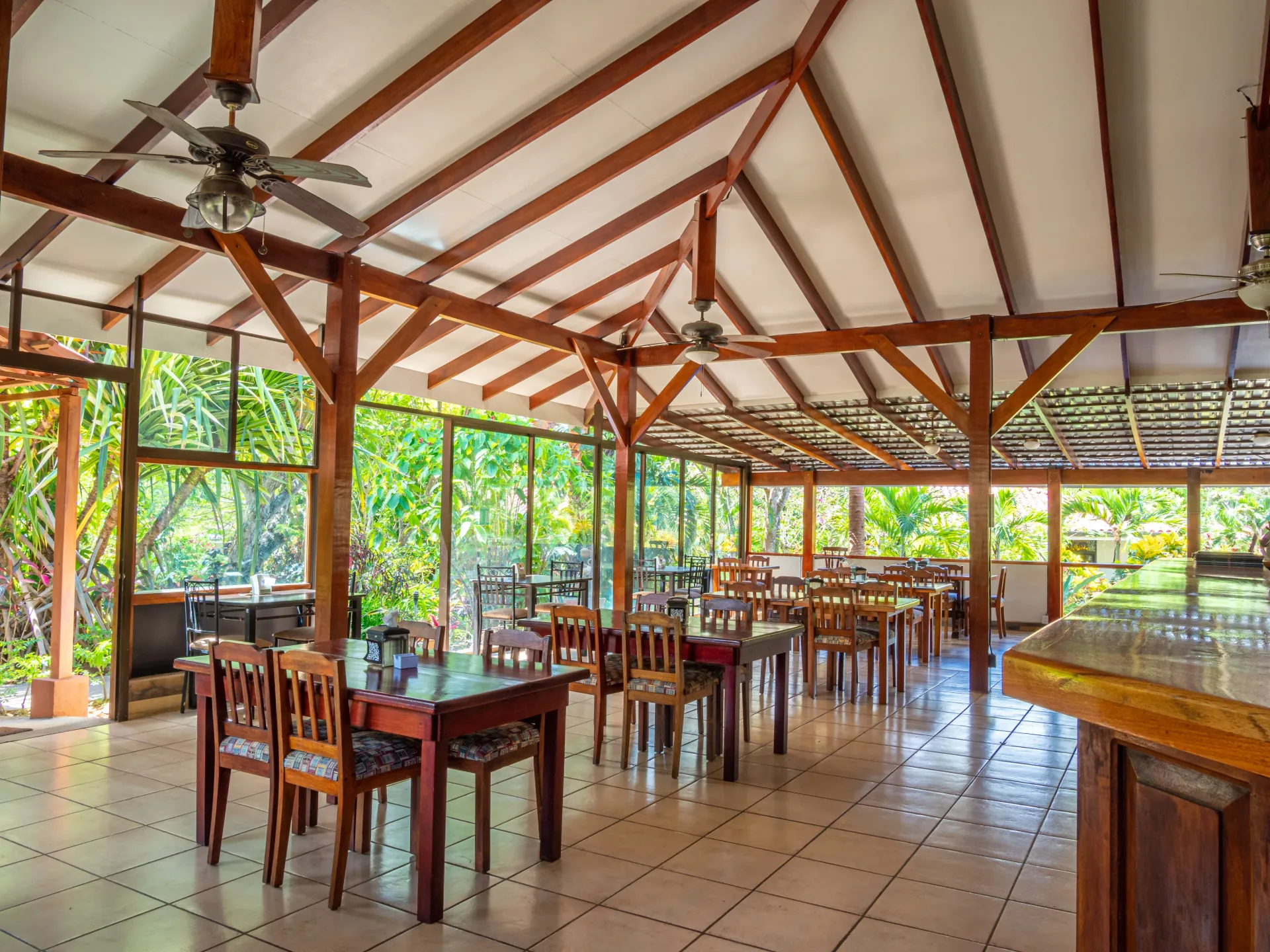Open-air restaurant with wooden tables and chairs under a high timber roof, surrounded by greenery.