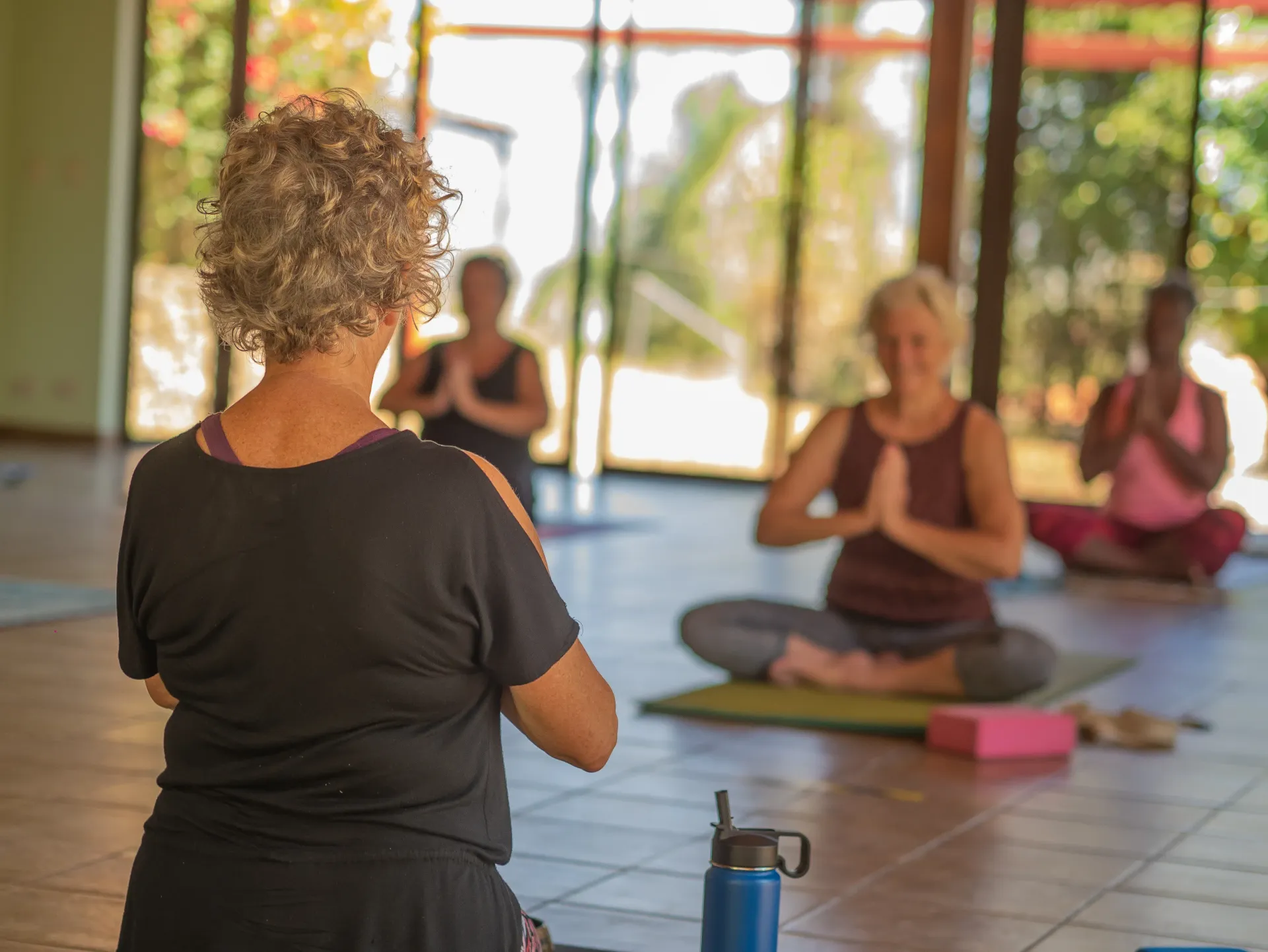 Yoga class in a sunlit studio, with participants seated on mats facing an instructor.