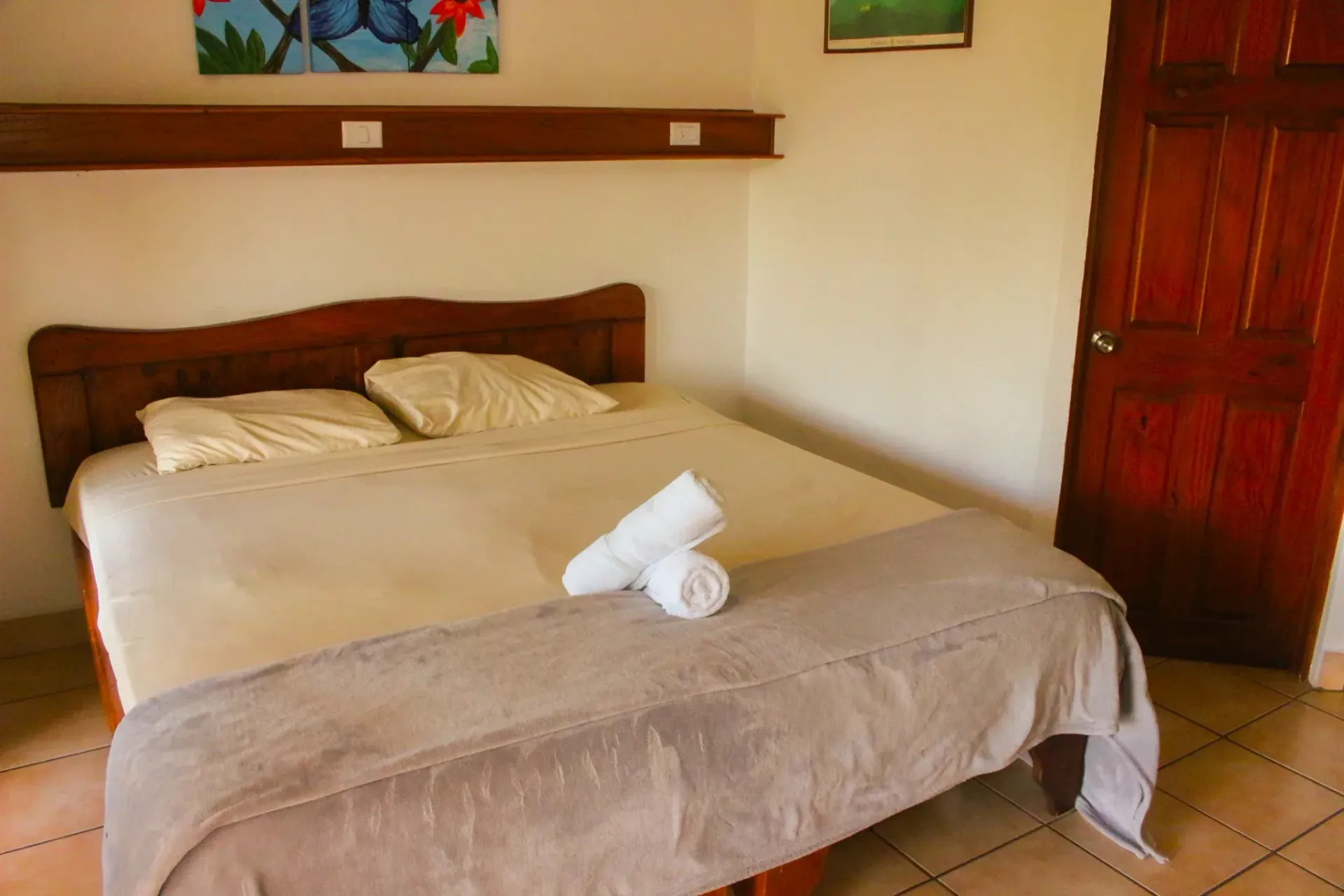 Habitación de hotel ordenada con cama doble, ropa de cama beige, toalla sobre la cama, estante superior y puerta de madera.