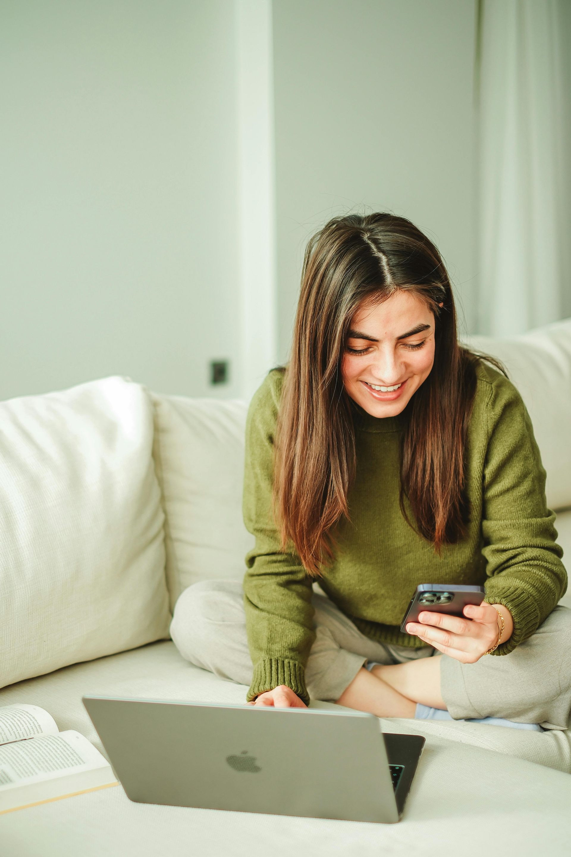 Una donna è seduta su un divano e usa un computer portatile e un telefono cellulare.