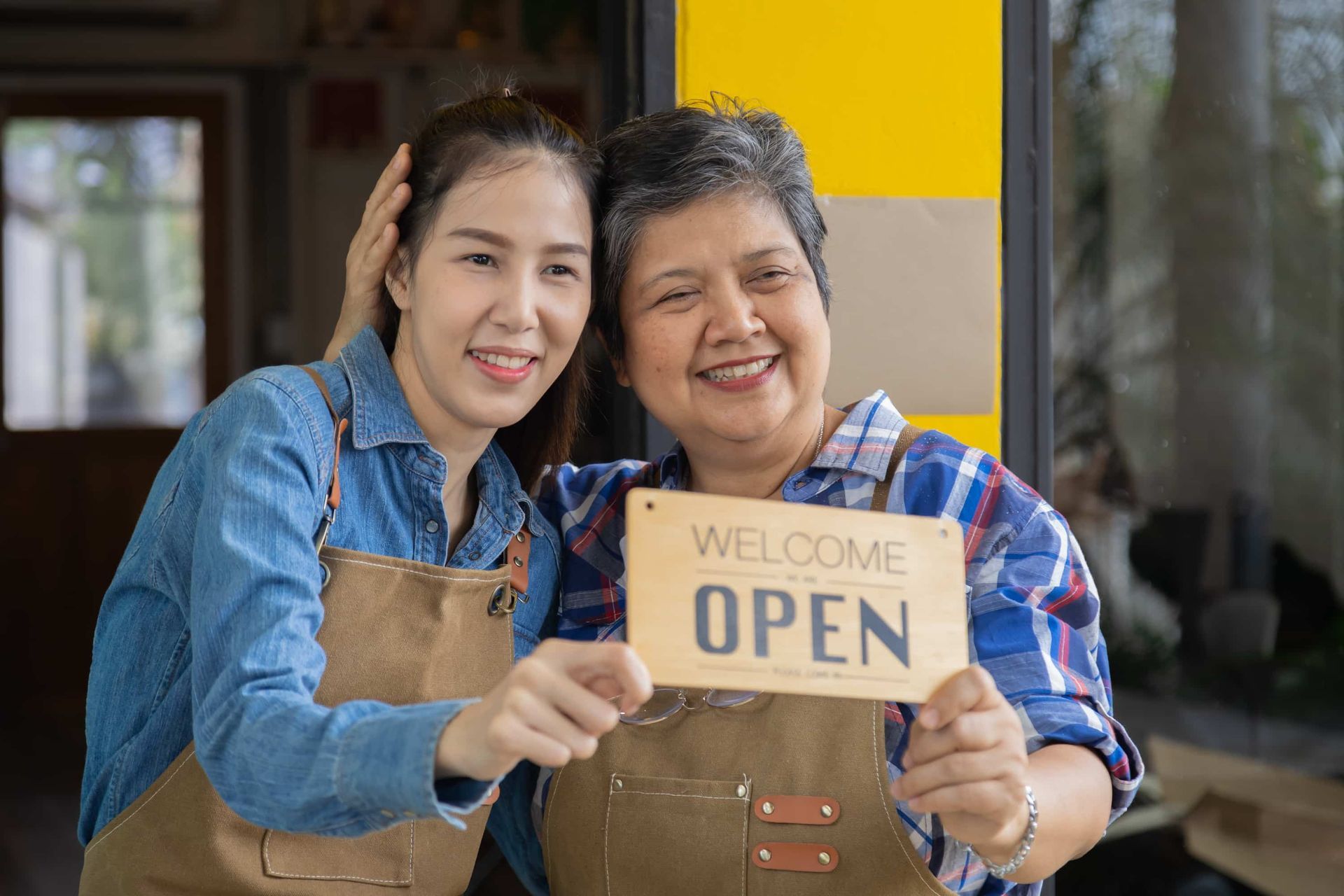 Two people smiling and holding a “Welcome Open” sign outside a storefront.