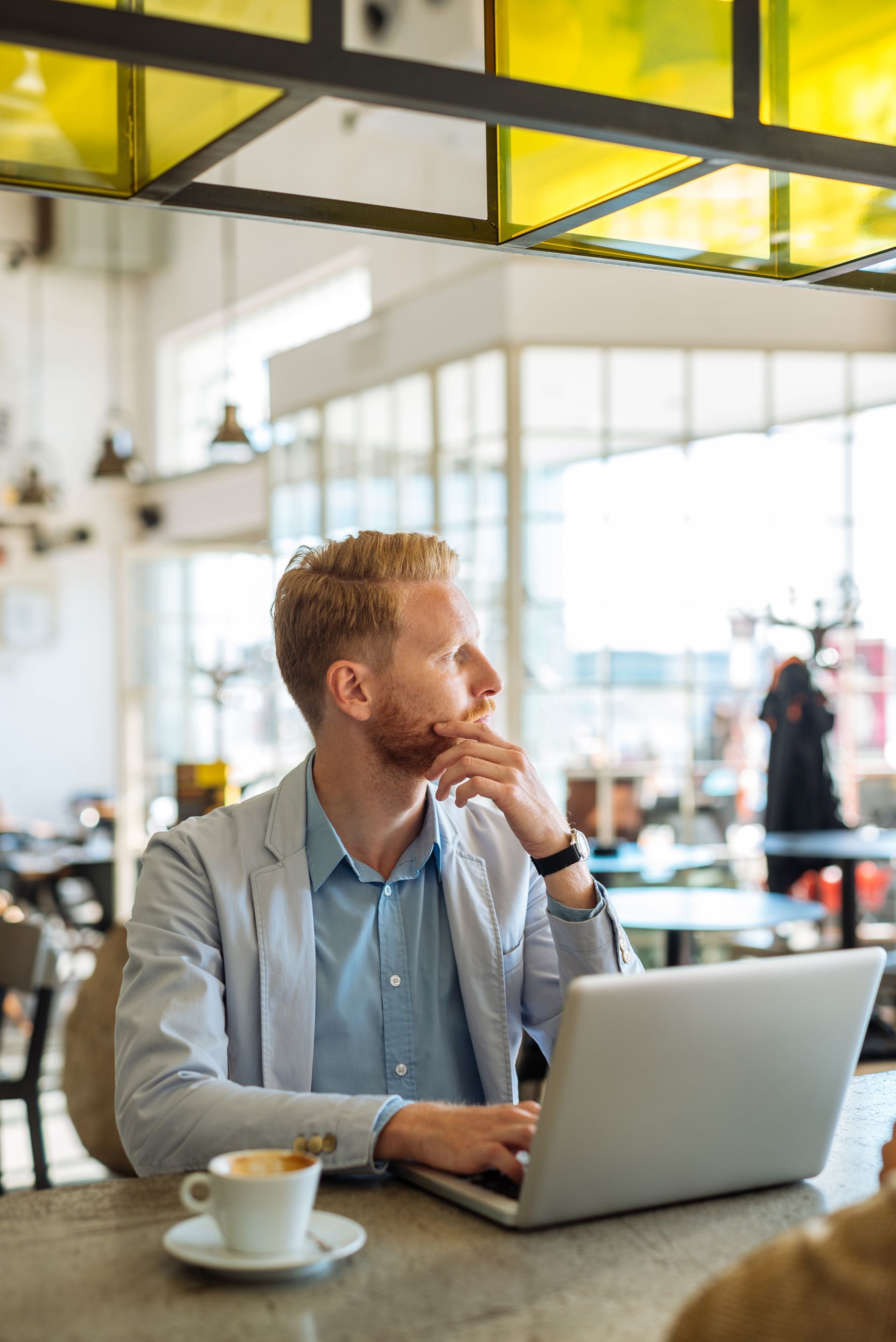 Man at a cafe, using laptop, looking thoughtful with hand on chin, near a coffee cup.