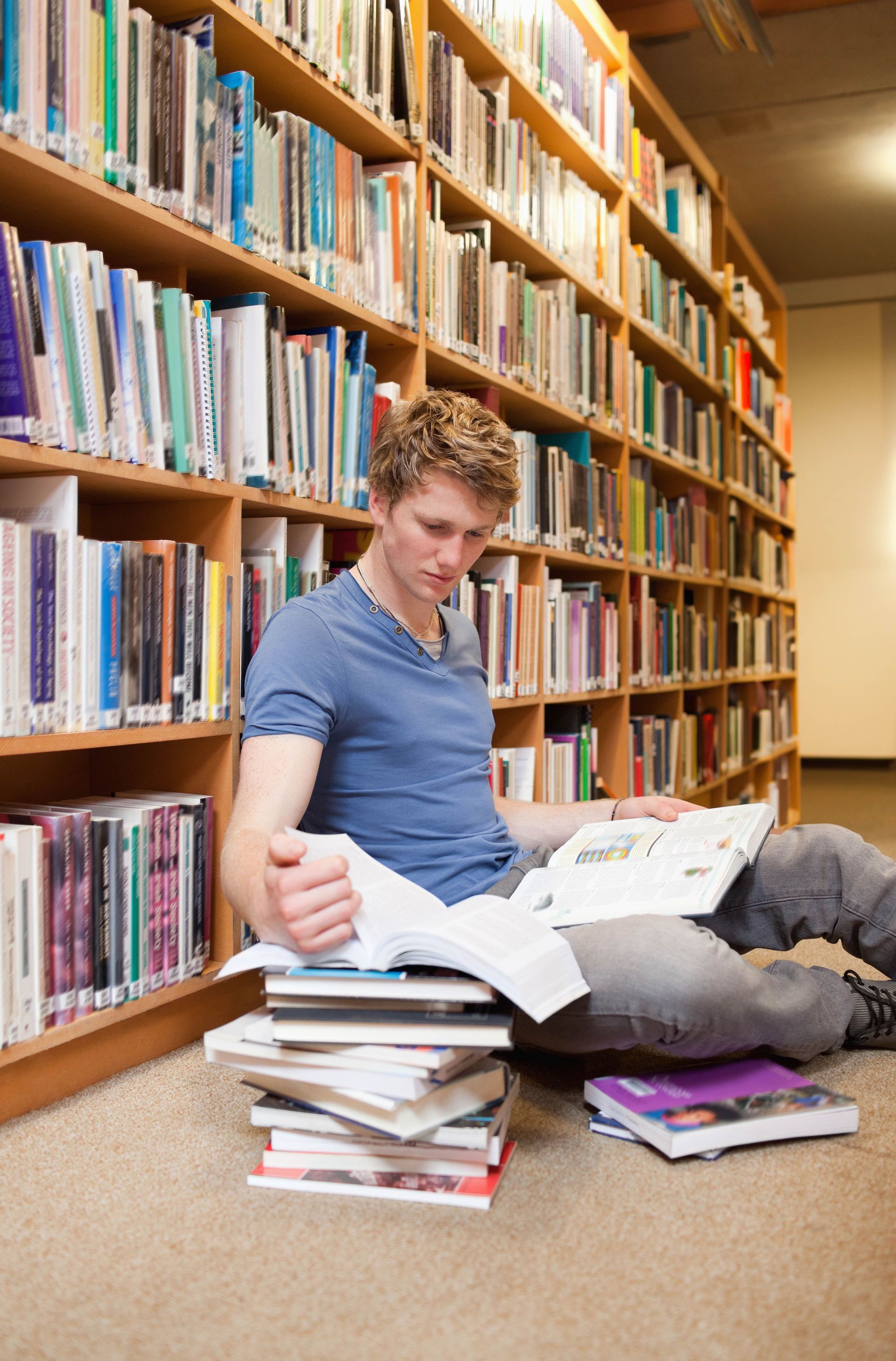 Person reading books on library floor, surrounded by bookshelves.