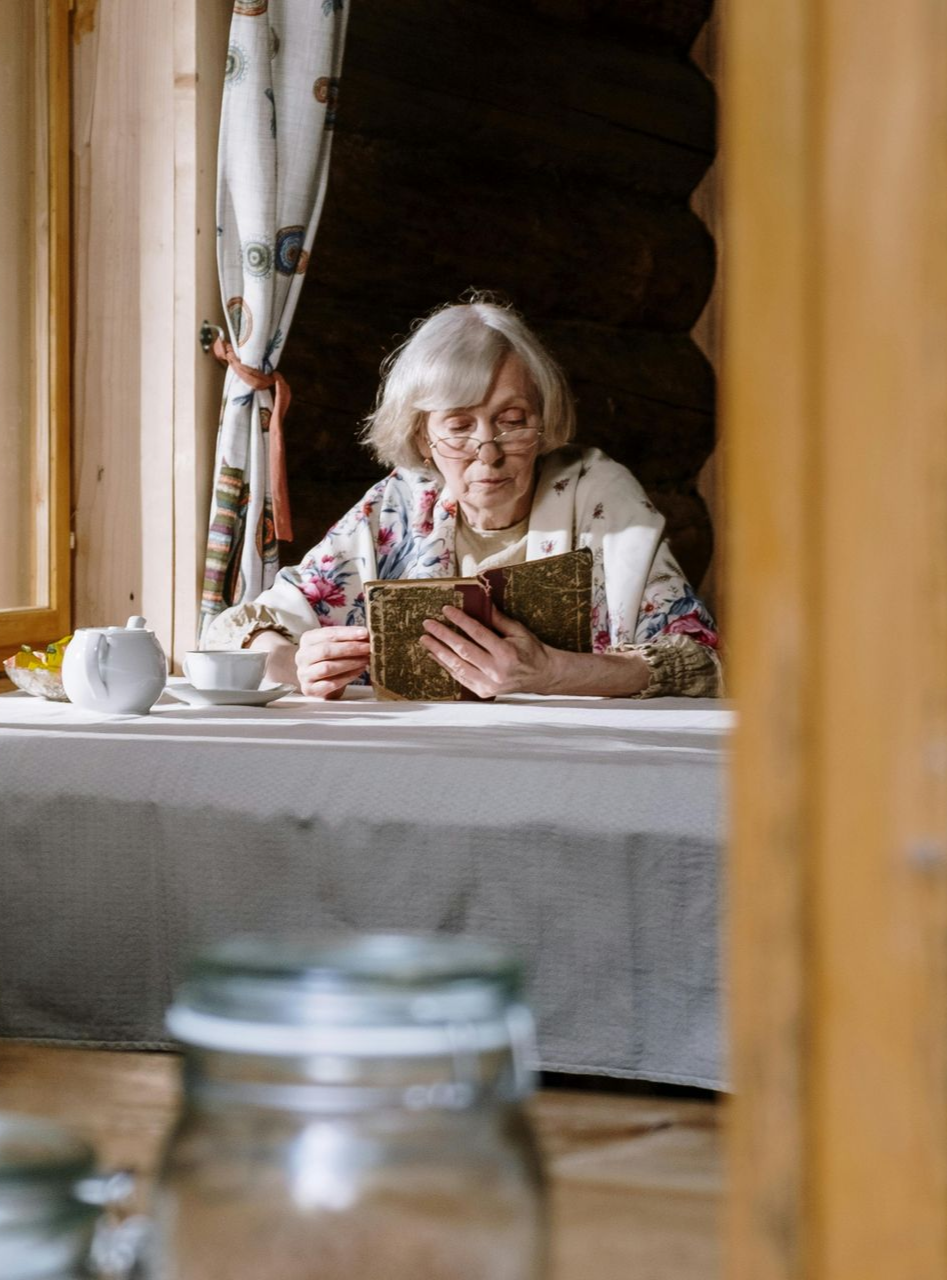 Elderly person reading a book at a table by a window, warm sunlight, tea set.