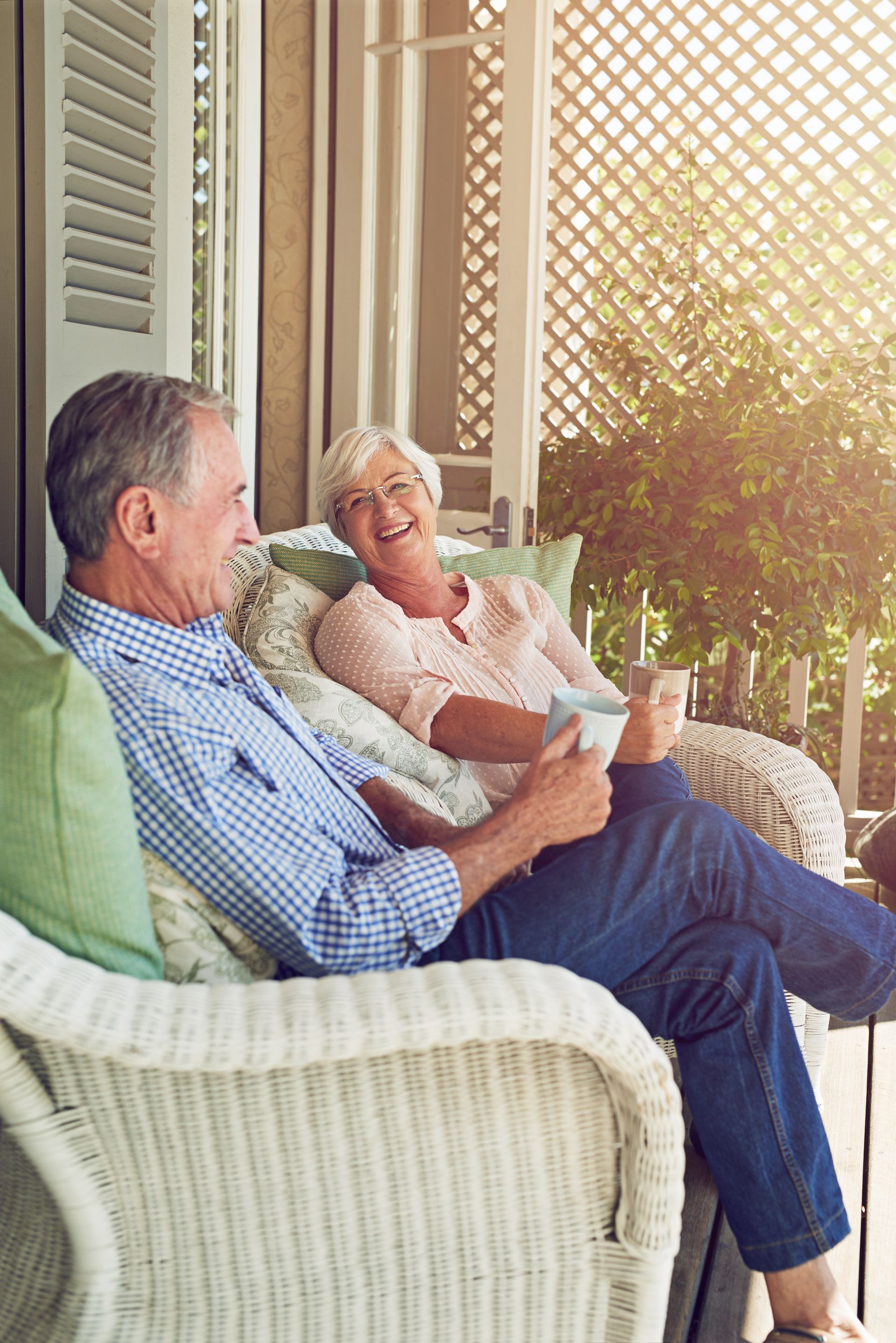 Senior couple laughing on a sunny porch, sitting in wicker chairs.