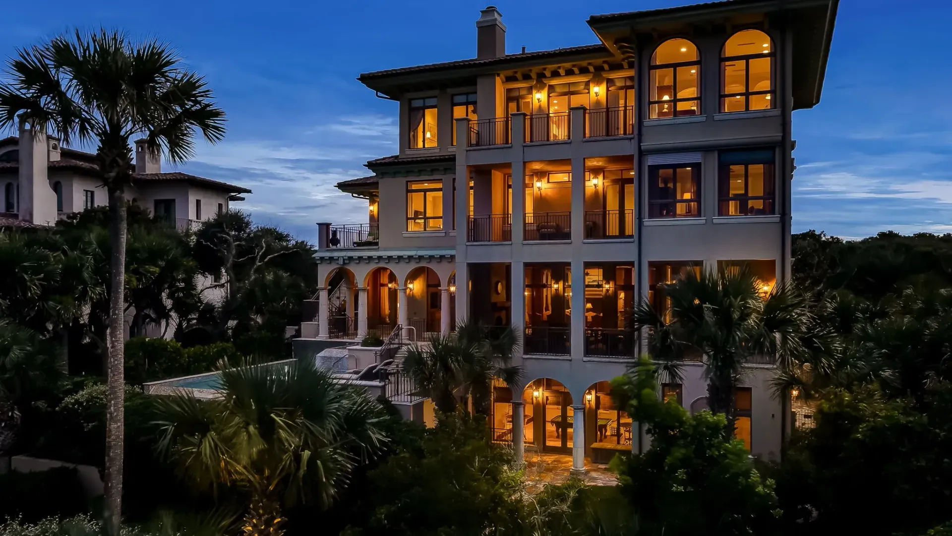 Multi-story luxury home at dusk, illuminated interior, surrounded by trees.