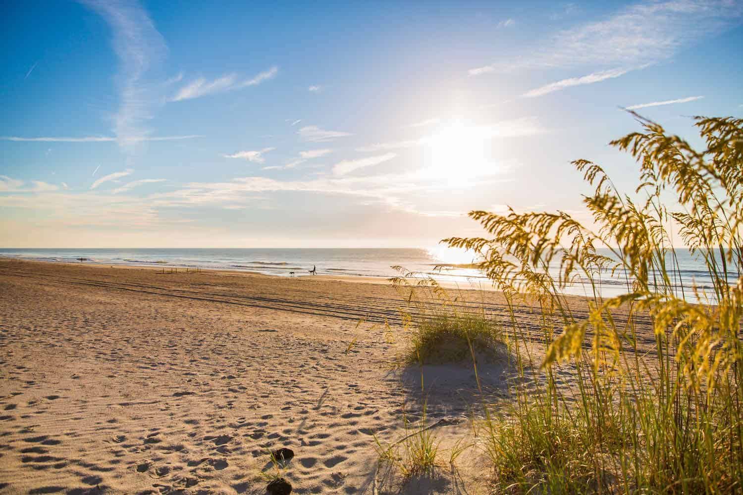 Sandy beach with sun shining over ocean waves and grassy dunes.