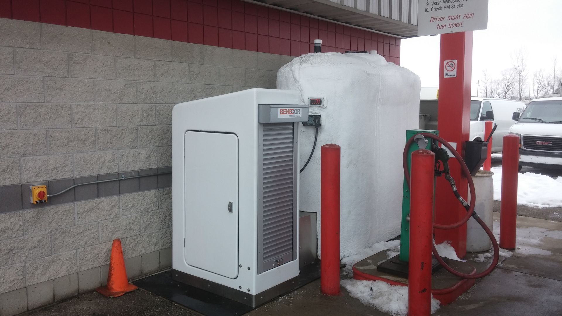 Gas pump with white unit and tank covered in snow, red poles, brick wall, snow covered ground.