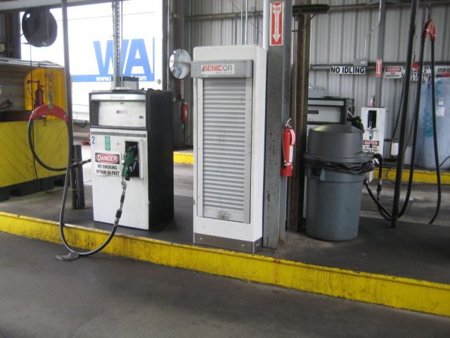 Fuel pump next to a utility cabinet in a garage. Yellow curb, fire extinguisher, trash can present.