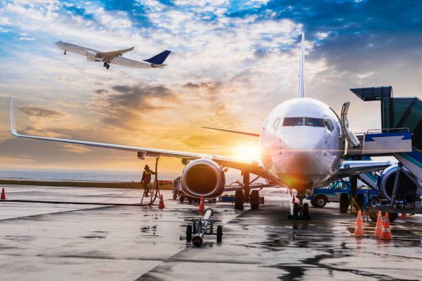 Airplane on tarmac with open door, another taking off, sunset sky.