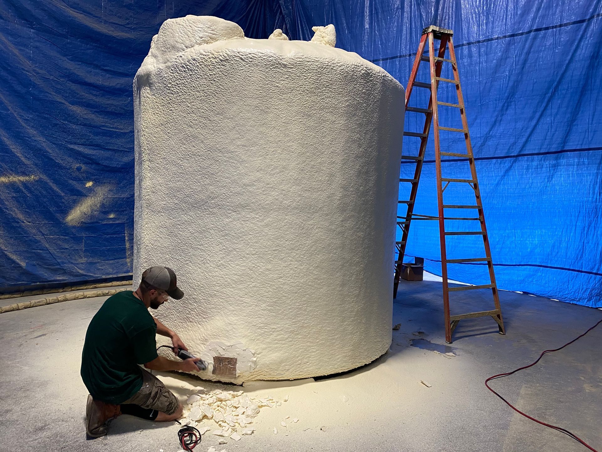 Man working on large, foam-covered tank in a blue-walled room; a ladder leans against the tank.