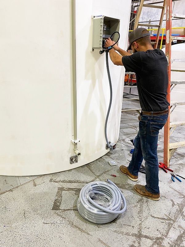 Man in jeans and cap wiring electrical box on a curved white structure; coiled gray cable on the floor.