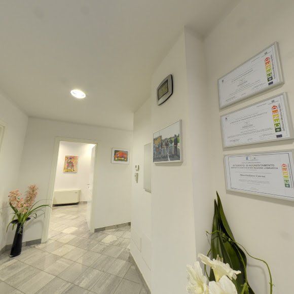 Luminoso corridoio d'ufficio bianco con pavimento piastrellato, opere d'arte alle pareti, certificati incorniciati e una pianta in vaso.