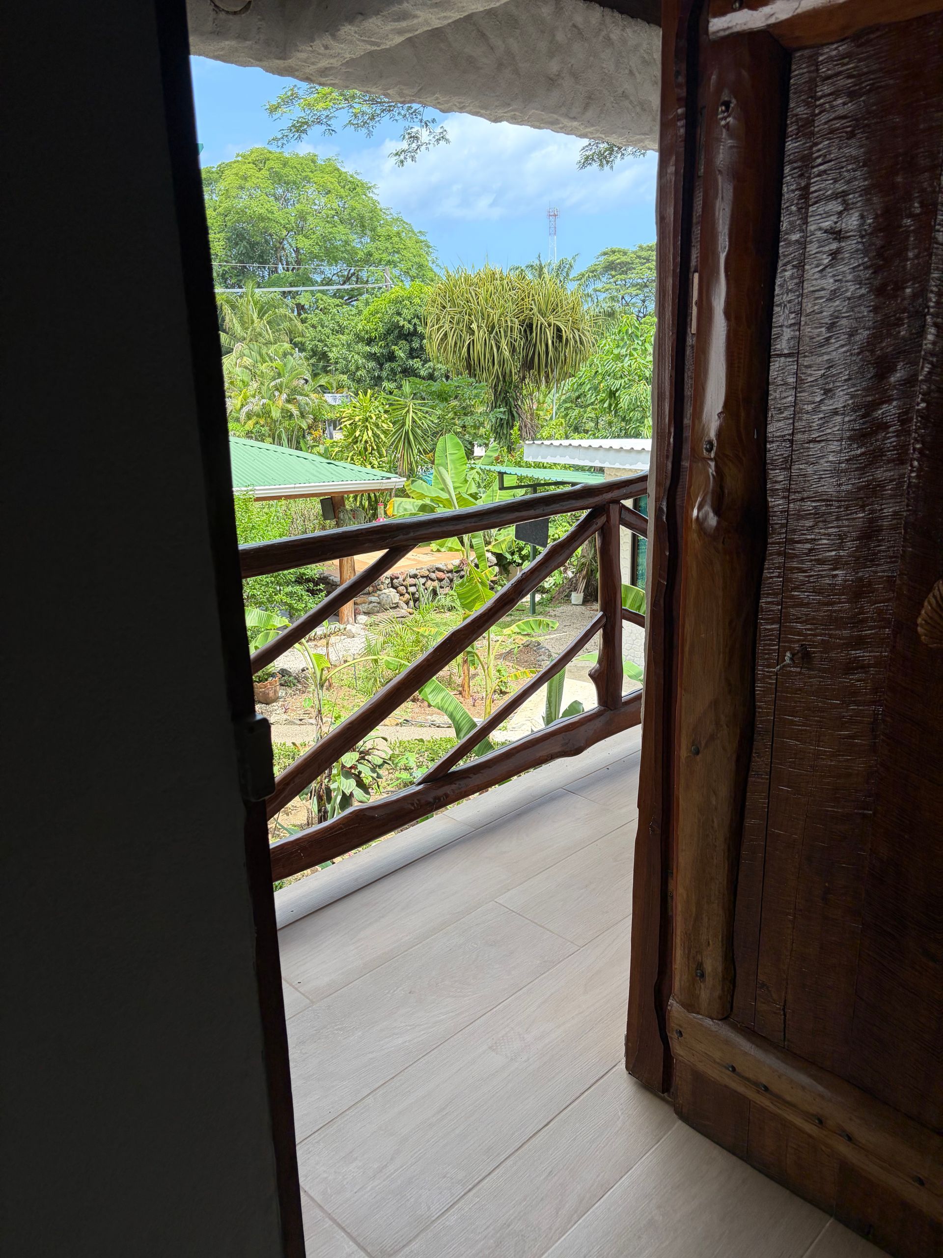 View from a doorway to a balcony with wooden railing and lush green tropical trees beyond.