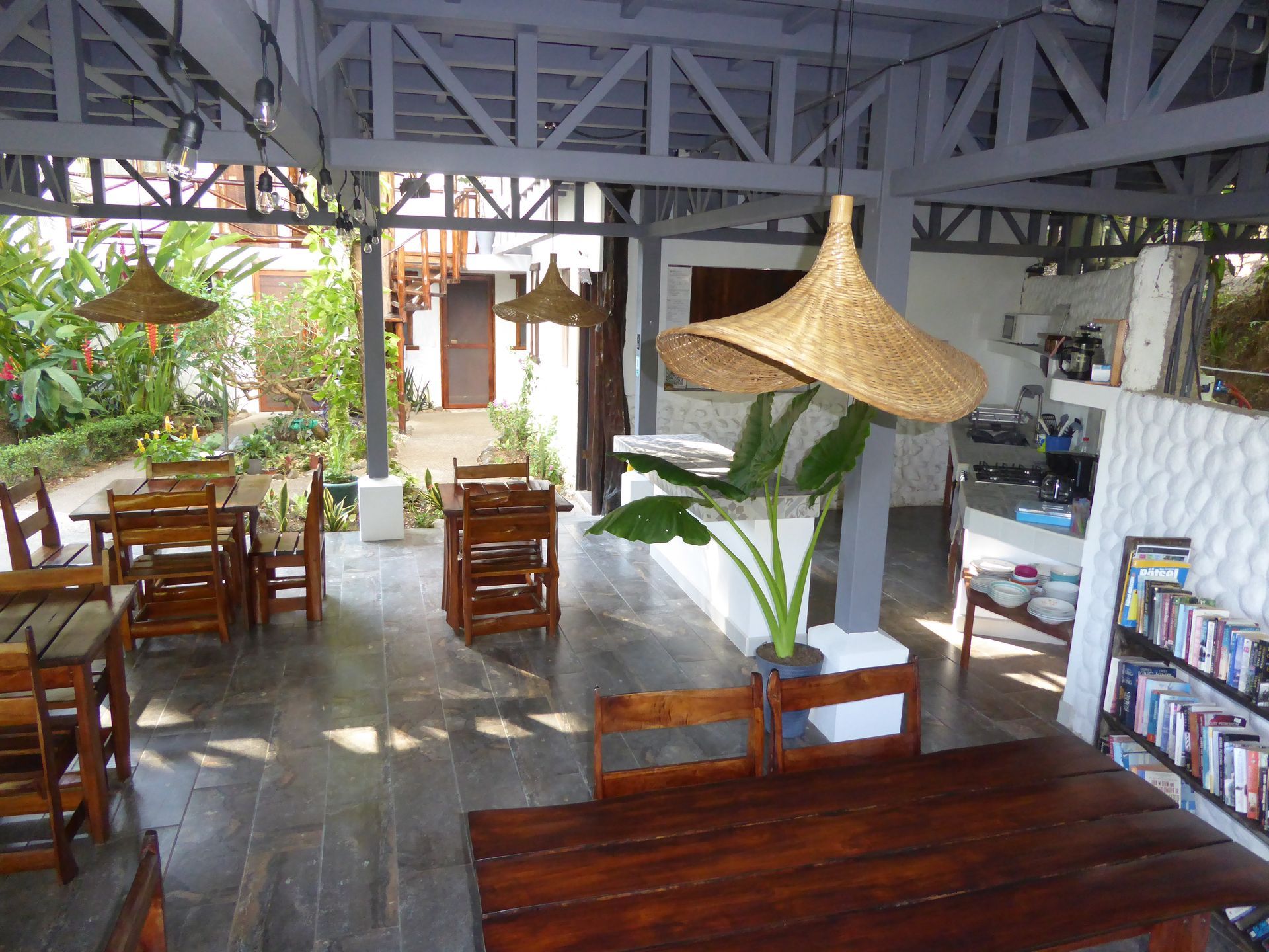 Indoor-outdoor cafe with wooden tables, chairs, and woven lampshades; greenery visible outside.