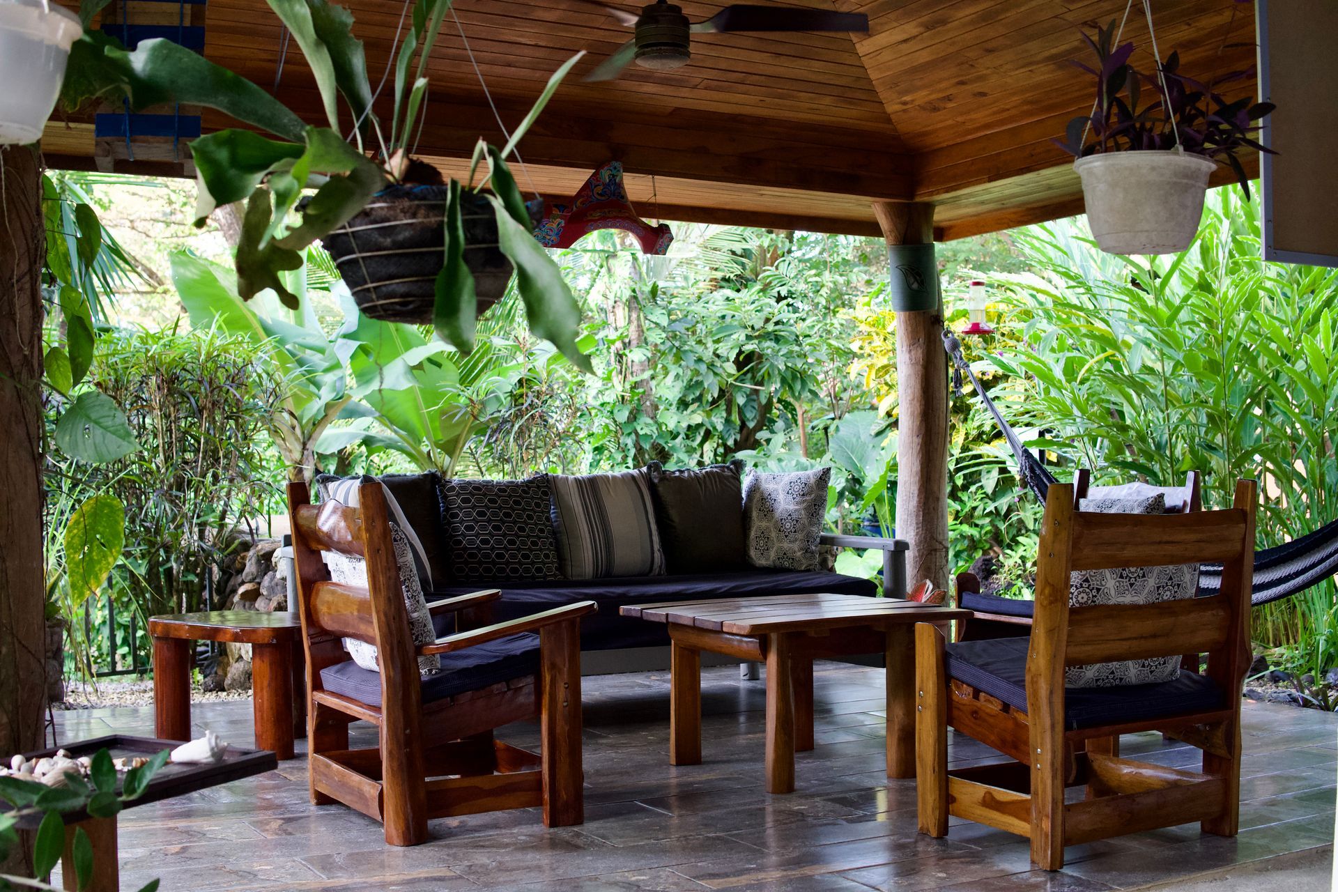 Outdoor seating area with wooden furniture, surrounded by lush greenery, under a thatched roof.