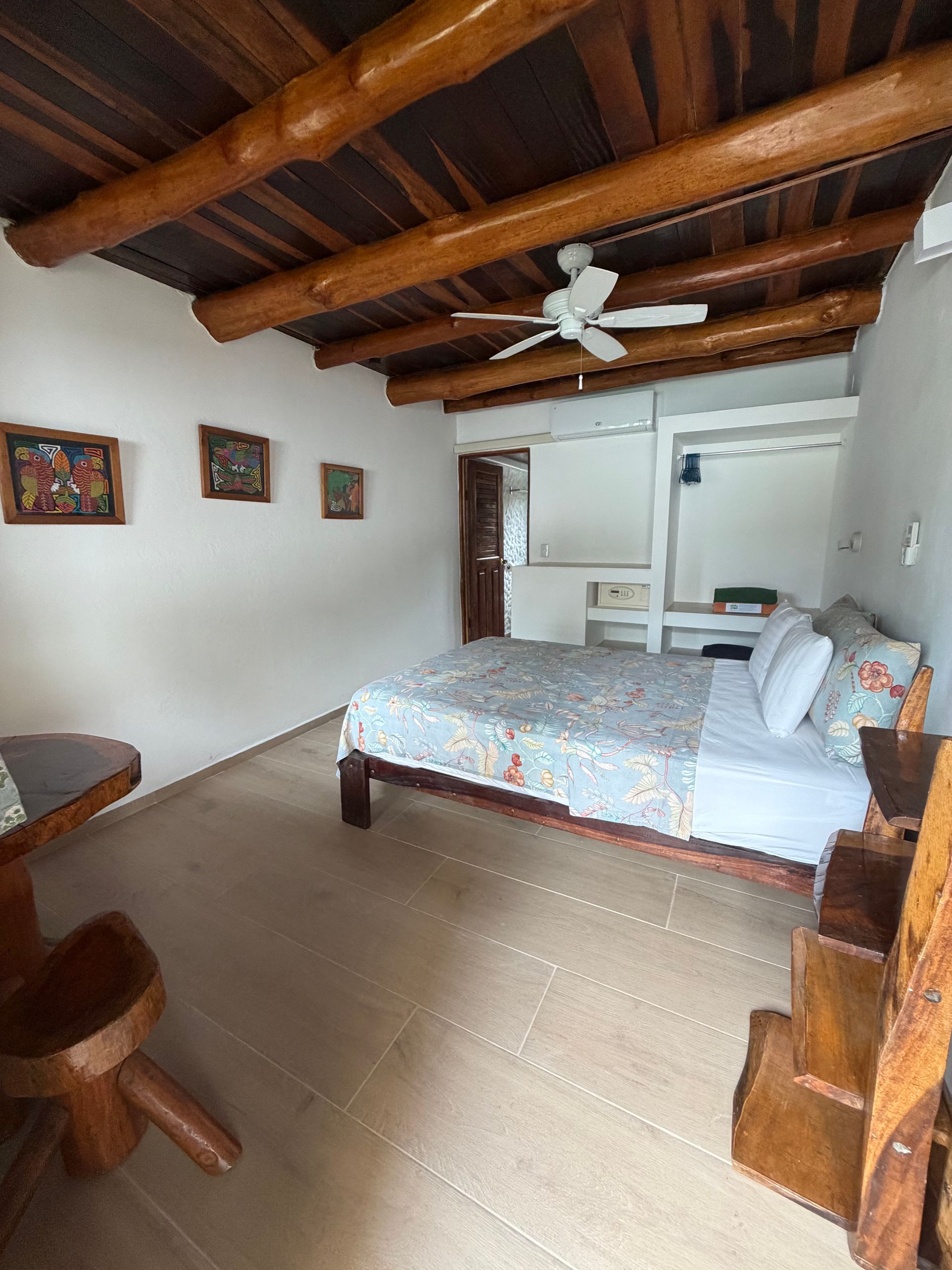 Rustic bedroom with exposed wooden beams, white walls, bed, ceiling fan, and small desk by the window.