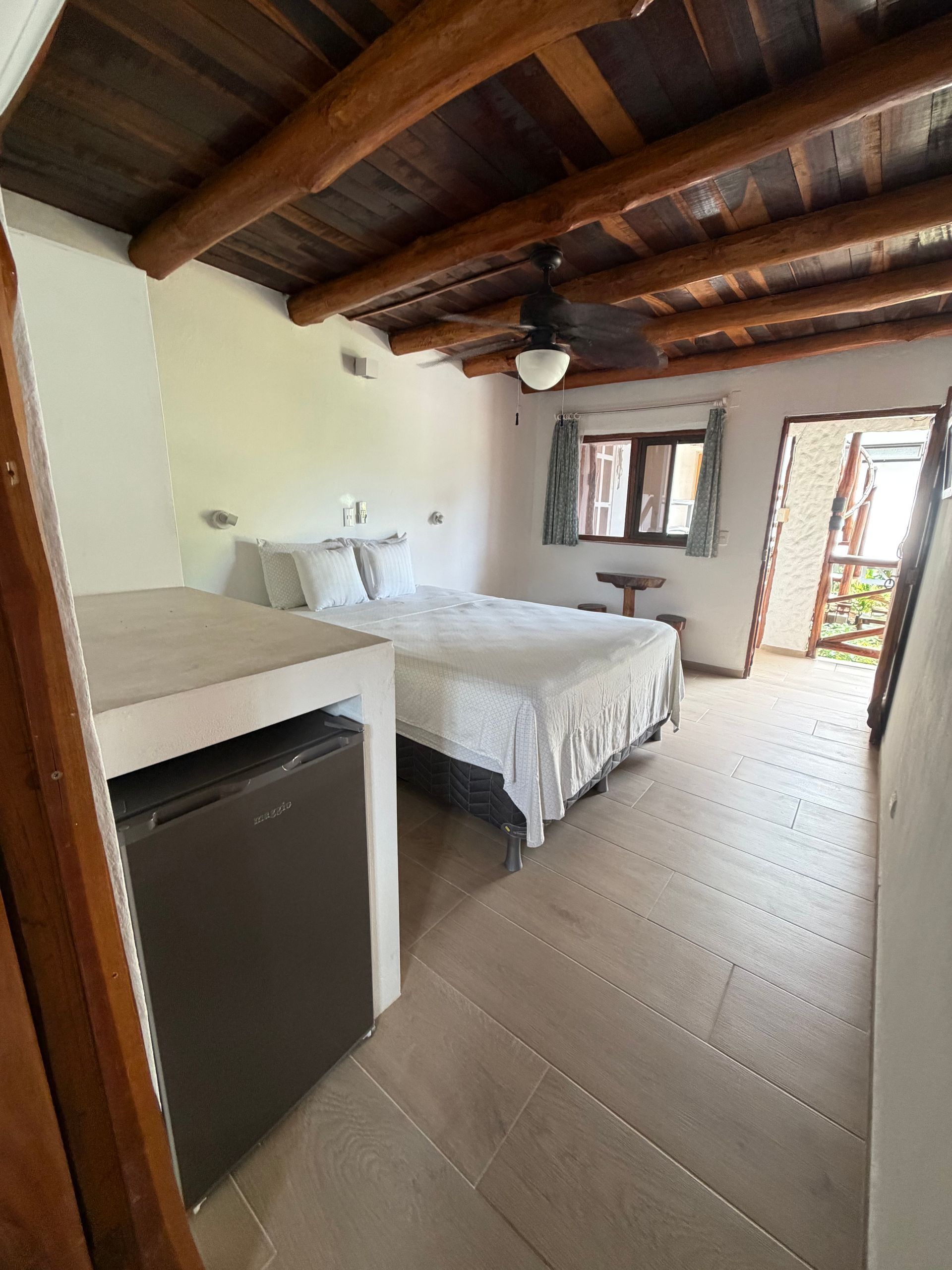 Small rustic bedroom with two beds, wood ceiling, window, and open door to a bright patio