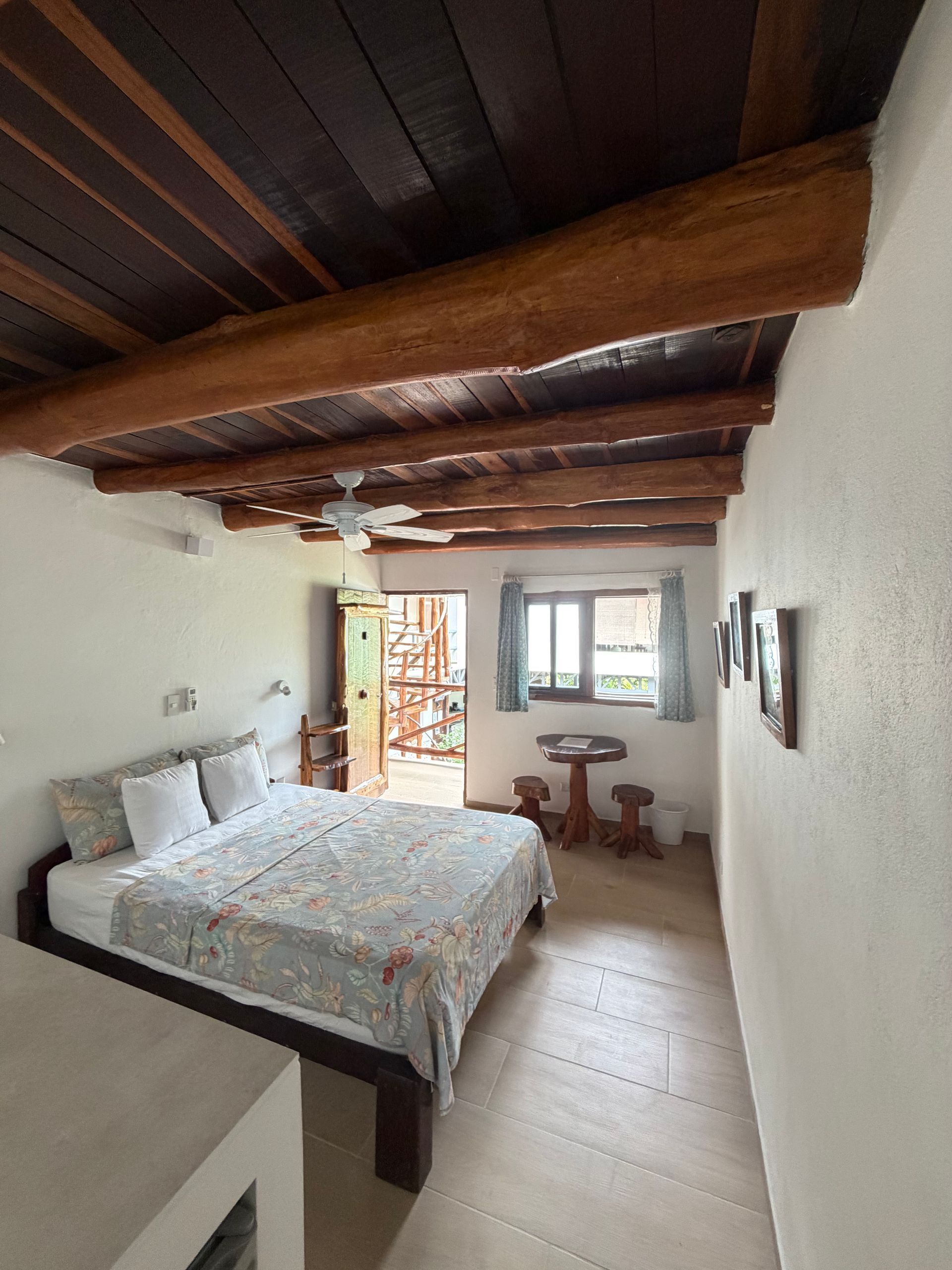 Small rustic bedroom with a bed, wood-beam ceiling, window, and open door to a sunny terrace